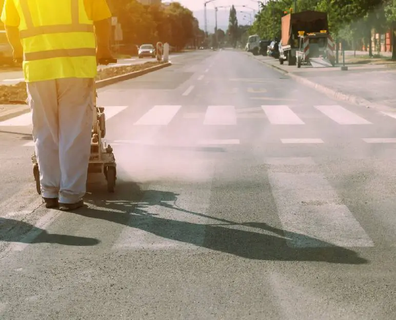 Worker in a yellow vest paints road lines on a sunny street